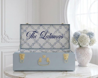 May include: A light blue storage trunk with gold-toned metal latches and a central handle. The interior lid features a white background with a blue floral trellis pattern and the text The Lalamas in a dark blue cursive script font.