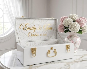 May include: A white rectangular keepsake box with gold-toned metal latches and a handle. The interior lid features a white floral pattern with the gold text "Emily & Tom" and "October 16, 2026" printed in a cursive script font.