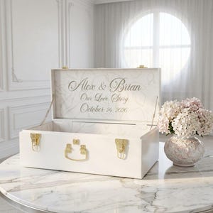 May include: A white rectangular wooden keepsake box with gold-toned metal latches and a central handle. The interior lid features the engraved text Alex & Brian, Our Love Story, October 24, 2026, set against a faint, light-colored floral pattern.