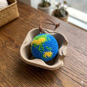 May include: A blue, yellow, and green crocheted globe ornament with a brown leather loop for hanging. The globe is displayed in a tan, paper-like container. The continents are represented in yellow and green against a blue ocean.