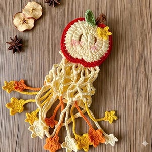 May include: A handmade crochet apple-shaped decoration with a red border, cream-colored center, and green leaf. The apple has a yellow net-like design with star-shaped accents in yellow, orange, and cream colors. The craft is placed on a wooden surface.