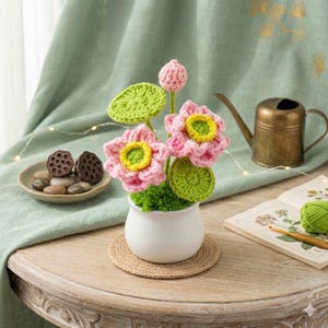 Puede incluir: Un arreglo floral de ganchillo hecho a mano con flores de loto rosas con centros amarillos y hojas verdes, en una maceta de cerámica blanca. El arreglo se encuentra sobre un posavasos de yute, con una regadera y un libro abierto en el fondo.
