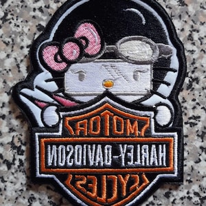 May include: Embroidered patch featuring a stylized Hello Kitty character wearing a black helmet and goggles. The patch includes a pink bow and the Harley-Davidson logo in orange and white.