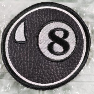 Puede incluir: Parche bordado en blanco y negro con el número 8 dentro de un círculo. El número 8 es blanco, y el fondo es negro con una apariencia texturizada. Un borde blanco rodea el diseño.