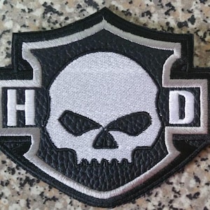 Könnte beinhalten: Schwarz-weißer, gestickter Aufnäher mit einem Totenkopf-Design in Schildform. Die Buchstaben "H" und "D" befinden sich auf beiden Seiten des Schädels. Der Aufnäher hat einen silbernen Rand und einen strukturierten schwarzen Hintergrund.