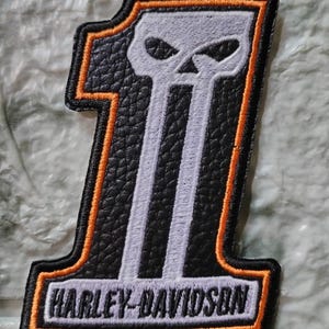 May include: A black and orange Harley-Davidson patch in the shape of the number one, with a skull design and the brand name. The patch has a textured leather-like appearance and is outlined in orange.