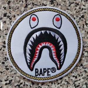 May include: A circular white patch featuring a shark face design with red eyes, a black open mouth with white teeth, and red accents. The patch is bordered by a gold rope design and has the word "BAPE®" in black.
