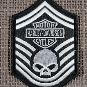 Puede incluir: Parche Harley-Davidson en blanco y negro con un diseño de calavera y huesos cruzados. El parche presenta el logotipo de Harley-Davidson y la palabra "CYCLES". El parche tiene un borde negro y rayas blancas.