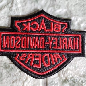 Puede incluir: Parche Harley-Davidson negro y rojo con las palabras "HARLEY-DAVIDSON" y "BLACK RIDERS" bordadas en rojo. El parche tiene forma de escudo con un borde negro y una apariencia texturizada.