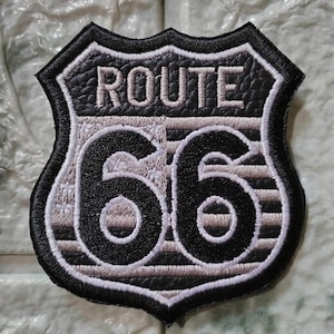 Puede incluir: Parche bordado de la Ruta 66 en blanco y negro. El parche tiene forma de escudo y presenta las palabras "ROUTE" sobre el número "66". El diseño tiene un aspecto texturizado y un borde blanco.