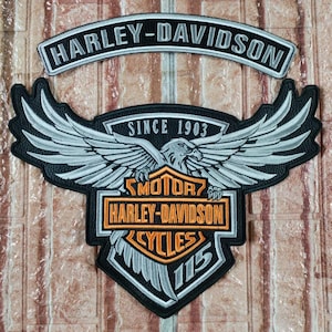 Puede incluir: Un parche de Harley-Davidson en blanco y negro con un diseño de águila y alas. El parche incluye el texto "HARLEY-DAVIDSON" y "CYCLES" con el texto "SINCE 1903" y "115".