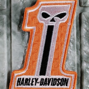 Puede incluir: Parche bordado con el número "1" en naranja, con un diseño de calavera y el logotipo de Harley-Davidson. El parche tiene una combinación de colores en blanco y negro, con el texto "HARLEY-DAVIDSON" en la parte inferior.