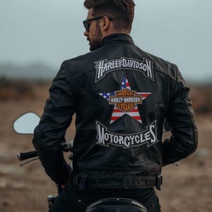 Puede incluir: Chaqueta de moto de cuero negro con el logo de Harley-Davidson en la espalda. El logo presenta una estrella con el diseño de la bandera estadounidense y las palabras "Motorcycles" y "Harley-Davidson".