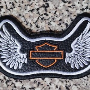 May include: Black patch featuring white embroidered wings flanking an orange and black emblem. The patch has a black border with a white outline. The emblem has an orange outline and a black interior.