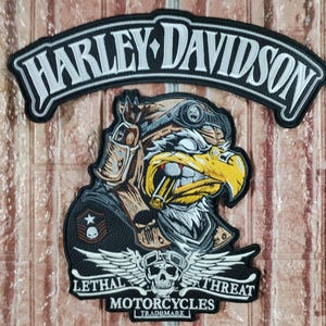 Pode incluir: Um patch Harley-Davidson em preto e branco com um gráfico de águia usando um capacete de piloto e segurando um charuto. O patch também apresenta as palavras "Lethal Threat Motorcycles" e uma caveira com asas.