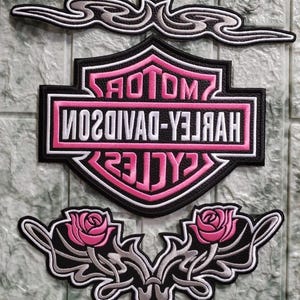 May include: Embroidered patches featuring the Harley-Davidson logo in black, pink, and white. The set includes the iconic shield logo, decorative swirls, and floral designs with roses. These patches are suitable for jackets, vests, or bags.