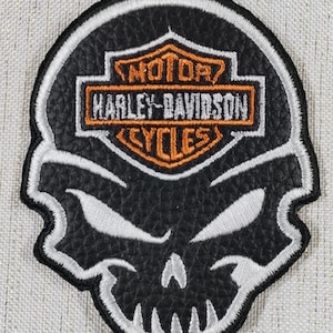 Puede incluir: Parche negro y blanco de Harley-Davidson Cycles con forma de calavera. El parche presenta el logotipo de Harley-Davidson en naranja y blanco, con las palabras "MOTOR HARLEY-DAVIDSON CYCLES". La calavera tiene ojos y dientes bordados en blanco.