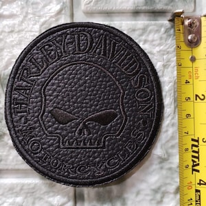 May include: A round, black Harley-Davidson patch with a skull design. The patch is approximately 3 inches in diameter. The text "HARLEY-DAVIDSON MOTORCYCLES" is embroidered around the skull.