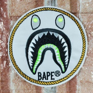 May include: A circular patch with a shark-themed design. It has a white background, black mouth, and green highlights. The patch is edged with a gold rope-style border and features the text "BAPE®" at the bottom.