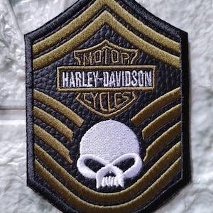 May include: Black and gold Harley-Davidson patch with a skull and the brand's logo. The patch has a textured background and a stitched border. The text "MOTOR HARLEY-DAVIDSON CYCLES" is embroidered on the patch.