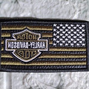 Puede incluir: Parche bordado con el logotipo de Harley-Davidson y un diseño de bandera estadounidense. El parche tiene un borde negro, rayas doradas y negras, estrellas blancas y el logotipo de Harley-Davidson en dorado y blanco. El texto "HARLEY-DAVIDSON" es visible.