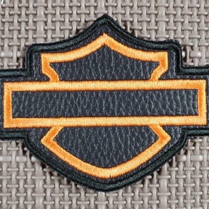Op de afbeelding: Een zwart en oranje geborduurd embleem in schildvorm. De patch heeft een zwarte achtergrond met een oranje rand en stiksels. Het ontwerp omvat een horizontale rechthoek en een schildvorm.