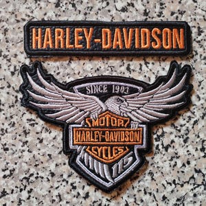 Puede incluir: Dos parches bordados de Harley-Davidson. El parche superior es rectangular con letras naranjas "HARLEY-DAVIDSON". El parche inferior presenta un diseño de águila alada, el texto "SINCE 1903" y el logotipo de Harley-Davidson.