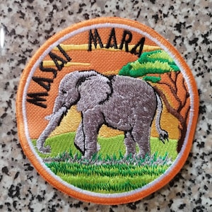 Könnte beinhalten: Gestickter Aufnäher mit einem grauen Elefanten, der auf grünem Gras geht, mit einem Baum und dem Text "MASAI MARA" in Schwarz. Der Aufnäher hat einen orangefarbenen Hintergrund und Rand.