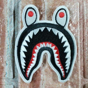 May include: Embroidered shark head patch. The patch has a black mouth with white teeth, red accents, and white outlines. The shark's head has two white ears with red circles. Ideal for clothing.