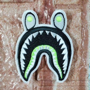 Puede incluir: Parche bordado con diseño de cabeza de tiburón estilizada. El parche tiene fondo negro, dientes blancos y boca interior verde. Las orejas son gris claro con un punto verde. Contorno blanco, ideal para ropa o accesorios.
