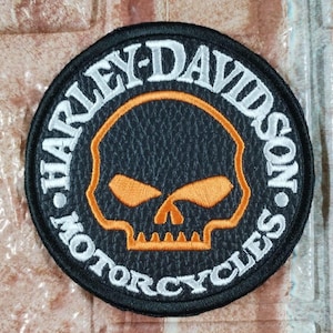 May include: A circular Harley-Davidson Motorcycles patch in black, with white embroidered text. The patch features an orange skull design. The words "HARLEY-DAVIDSON MOTORCYCLES" are embroidered around the skull.