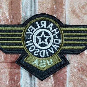 Puede incluir: Parche bordado con un diseño de alas en negro y verde oliva, con el texto "HARLEY-DAVIDSON USA" en un emblema circular. El parche tiene una estrella en el centro y es probablemente para una chaqueta o chaleco.