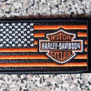 Puede incluir: Parche bordado con la bandera estadounidense en negro, blanco y naranja. El logotipo de Harley-Davidson está centrado, con las palabras "MOTOR HARLEY-DAVIDSON CYCLES" en un escudo. El parche tiene un borde negro.