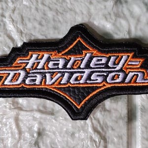 Può includere: Toppa Harley-Davidson ricamata in nero, arancione e bianco. La toppa presenta il logo Harley-Davidson in un design stilizzato con un bordo nero. La toppa è probabilmente destinata all'applicazione su abbigliamento o accessori.