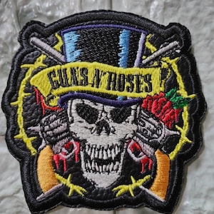 Puede incluir: Parche bordado con una calavera que lleva un sombrero de copa y sostiene dos revólveres. El logotipo de Guns N' Roses se muestra en una pancarta amarilla. El parche tiene un borde negro con alambre de púas amarillo.