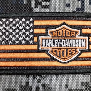 Puede incluir: Parche bordado con diseño de bandera americana, rayas naranjas y borde negro. Destaca el logotipo de Harley-Davidson Cycles. El parche es rectangular y tiene un borde negro.