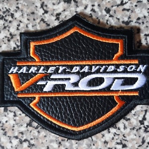 Puede incluir: Parche negro y naranja de Harley-Davidson V-Rod. El parche en forma de escudo presenta las palabras "HARLEY-DAVIDSON" y "V ROD" bordadas en hilo blanco y naranja. El parche tiene una apariencia texturizada similar al cuero.