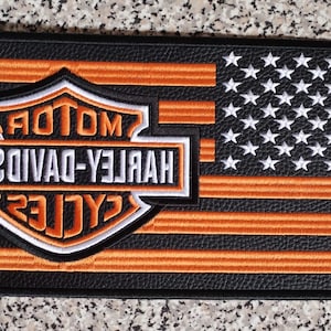 May include: A black and orange Harley-Davidson patch with an American flag design. The patch features the Harley-Davidson logo in the centre, with the words "MOTOR HARLEY-DAVIDSON CYCLES" in white. The flag has orange stripes and white stars.