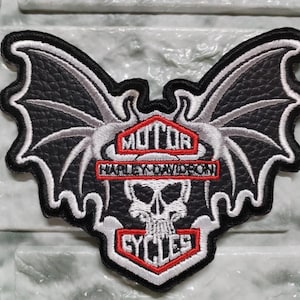 Peut inclure: Écusson brodé avec un motif de crâne ailé, intégrant le logo Harley-Davidson et le mot "CYCLES". Le patch est noir, blanc et rouge, avec un aspect texturé et des coutures blanches.
