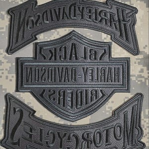 Puede incluir: Parches negros de motocicleta Harley-Davidson con letras en relieve. Los parches incluyen el logotipo de Harley-Davidson y las palabras "Black Riders" y "Motorcycles". Los parches están colocados sobre un fondo de camuflaje.