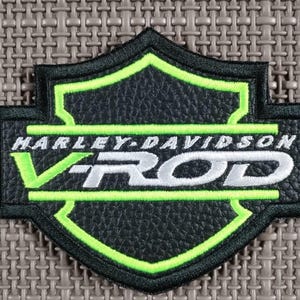 Puede incluir: Parche Harley-Davidson V-Rod negro y verde. El parche presenta el logotipo de Harley-Davidson con las palabras "HARLEY-DAVIDSON" y "V-ROD" en letras blancas. El parche tiene un borde negro y un contorno verde.