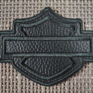 Puede incluir: Parche de cuero negro con forma de escudo y una barra rectangular en el centro. El parche tiene una superficie texturizada y un borde bordado en verde oscuro. El parche está sobre un fondo texturizado de color neutro.