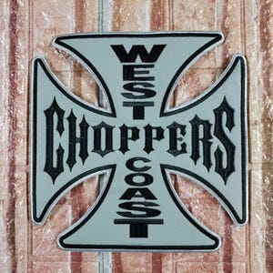 Puede incluir: Un parche gris en forma de cruz con un borde blanco. El parche presenta las palabras "WEST COAST CHOPPERS" en negro, con letras de estilo gótico. El parche es probablemente para un entusiasta de las motocicletas.