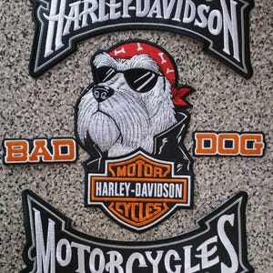 Puede incluir: Parches bordados con la marca Harley-Davidson y un bulldog con gafas de sol y un pañuelo rojo. Los parches incluyen las palabras "BAD DOG" y "MOTORCYCLES". Los parches son negros, blancos y naranjas.