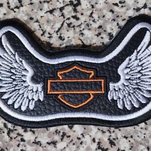 Puede incluir: Parche negro con alas bordadas en blanco y un emblema en forma de escudo con contorno naranja. El parche tiene un borde negro y una superficie texturizada. El emblema tiene un rectángulo naranja en el centro.
