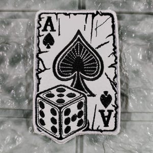May include: A white patch featuring a black Ace of Spades playing card design with a dice. The card has the letter "A" and the spade symbol. The dice shows the number six. The patch has a distressed border.
