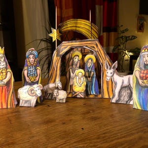 May include: A handmade nativity scene featuring paper cutouts of the Holy Family, the Three Wise Men, sheep, and a donkey. The figures are brightly colored with watercolor-style details, set against a backdrop of a star and a stable.