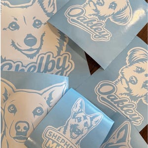 Puede incluir: Varias pegatinas de vinilo azul claro con ilustraciones de l&iacute;neas blancas de caras de perros. Los dise&ntilde;os incluyen los nombres Oakley y Shelby en letra cursiva, y una pegatina presenta el texto Shepherd Mix bajo un retrato de perro.
