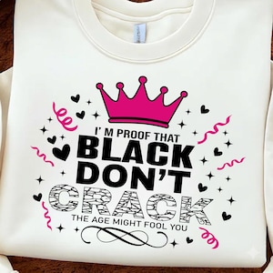 Puede incluir: Una sudadera de manga larga color crema con un diseño gráfico que incluye una corona rosa, el texto negro I'M PROOF THAT BLACK DON'T CRACK THE AGE MIGHT FOOL YOU, y espirales rosas decorativas, corazones negros y pequeñas estrellas.