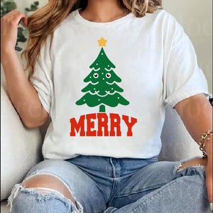 Op de afbeelding: Wit T-shirt met een groen kerstboomontwerp en een gele ster. Het woord "MERRY" is in het rood onder de boom gedrukt. Het shirt wordt gecombineerd met een blauwe spijkerbroek.